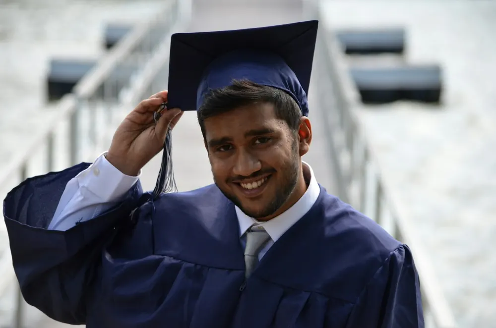 A smiling young man in a navy graduation gown, shirt and tie adjusts his mortarboard outdoors on a bright day