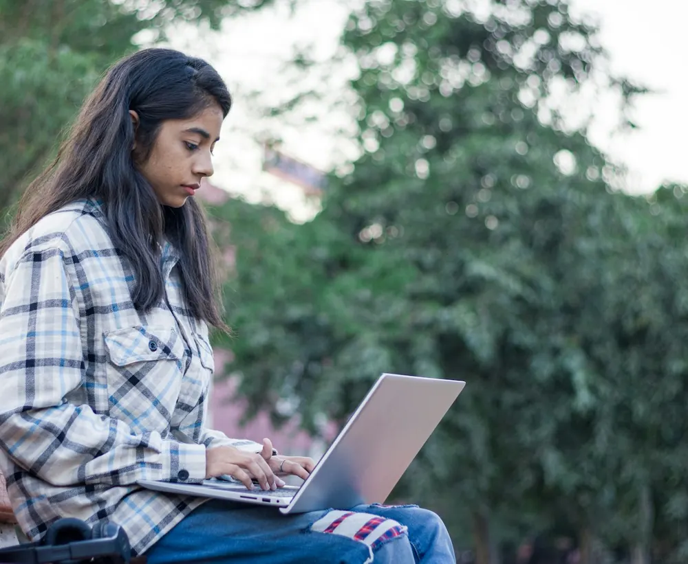 A young woman in a checked shirt types on a laptop while sitting on a park bench, with leafy green trees behind her