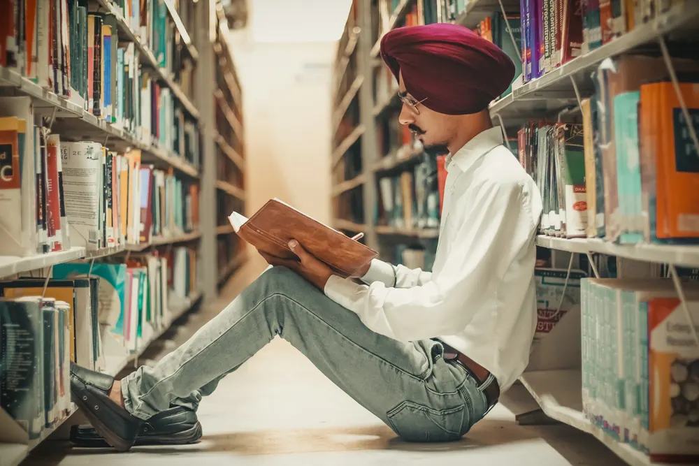 A young man in a maroon turban, glasses and a white shirt sits on a library floor between tall bookshelves, absorbed in a book