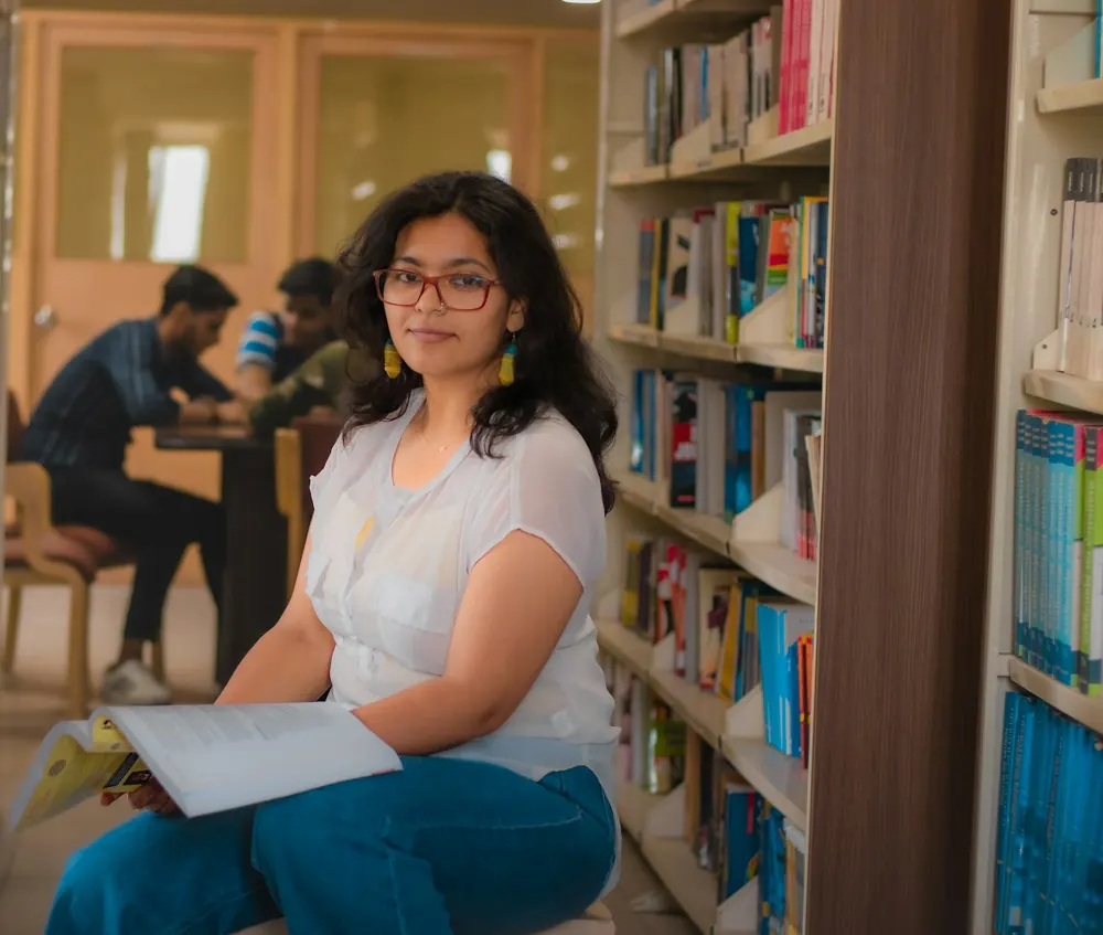A young woman in red glasses sits in a library aisle with an open book on her lap and smiles softly, while two students study at a table behind her