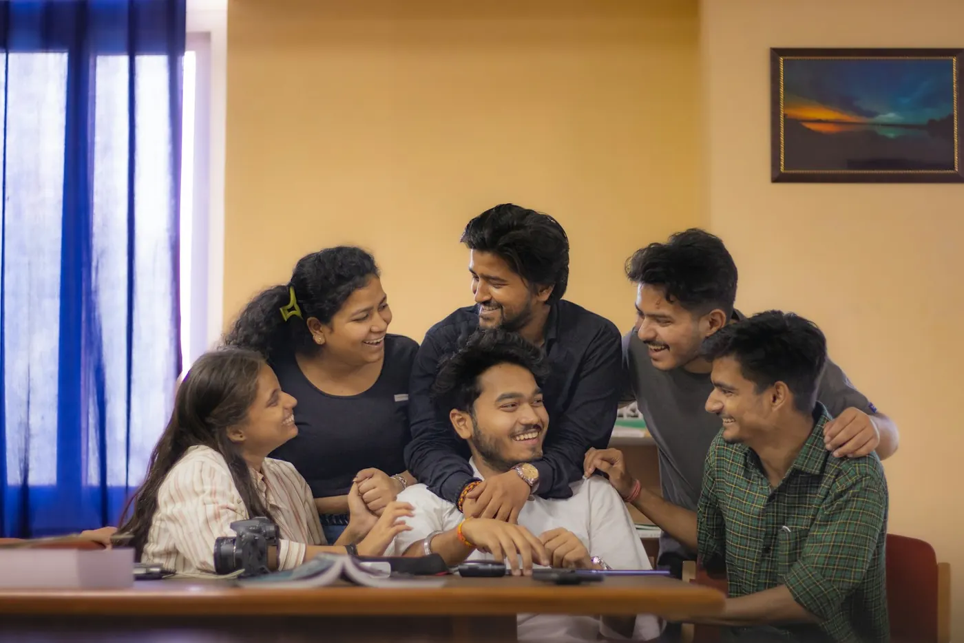 Six young students laugh together around a table with open books, in a sunlit room with a blue curtain and a warm yellow wall